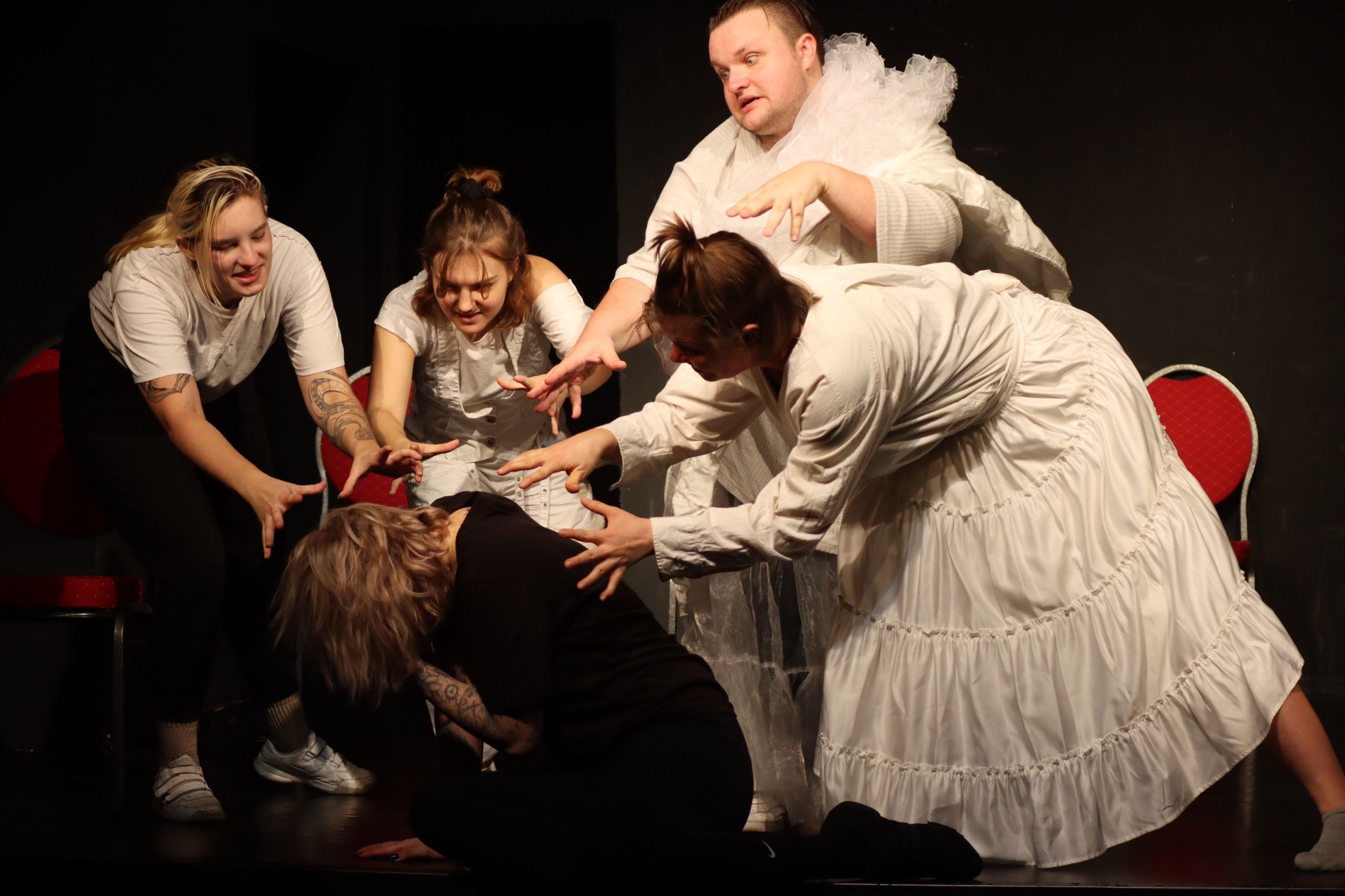 Szene auf der Bühne des Theaters Verlängertes Wohnzimmer: Eine Gruppe von Menschen in weißer Kleidung steht um eine auf den Boden gekauerte Frau in schwarzer Kleidung, die Hände nach vorne gestreckt, wie in einer Beschwörung